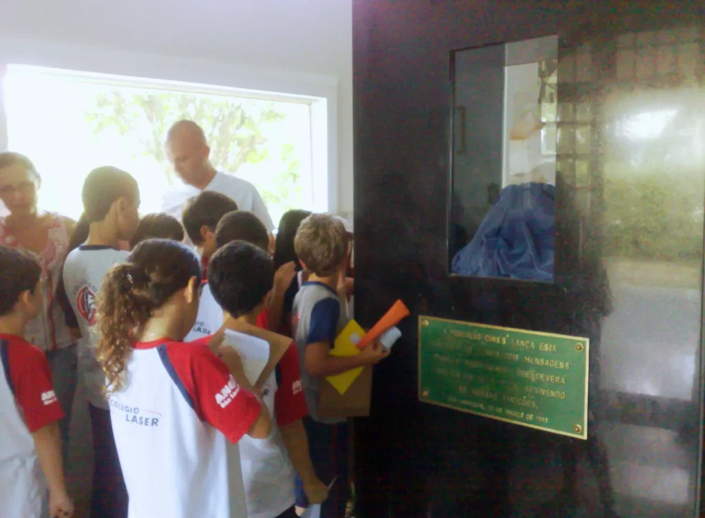 Alunos do colégio Laser visitando a Cápsula do Tempo na Fundação Cima’s. A imagem mostra um grupo de crianças e dois adultos observando a cápsula do tempo. A Cápsula é preta e tem uma câmara central de vidro, dentro da qual um cristal rosa transparente está posicionado sobre um pano azul. Há uma placa metálica verde com texto instalada na parte inferior da câmara de vidro. O grupo é composto por vários indivíduos vestindo roupas diferentes parecendo ser uniformes escolares; alguns estão segurando papéis ou pastas. Eles estão em um ambiente interno, com luz natural vindo de janelas ao fundo.
