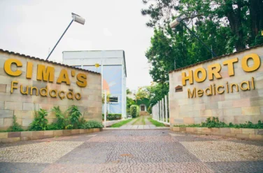 Entrada da Fundação Cima’s em São Lourenço, Minas Gerais, marcada por paredes de pedra com inscrições douradas que dizem: " Cima's Fundação" e " Horto Medicinal. Um caminho de paralelepípedos e vegetação exuberante, refletindo a rica história e legado do Professor Cima antes de seu falecimento em 2015.