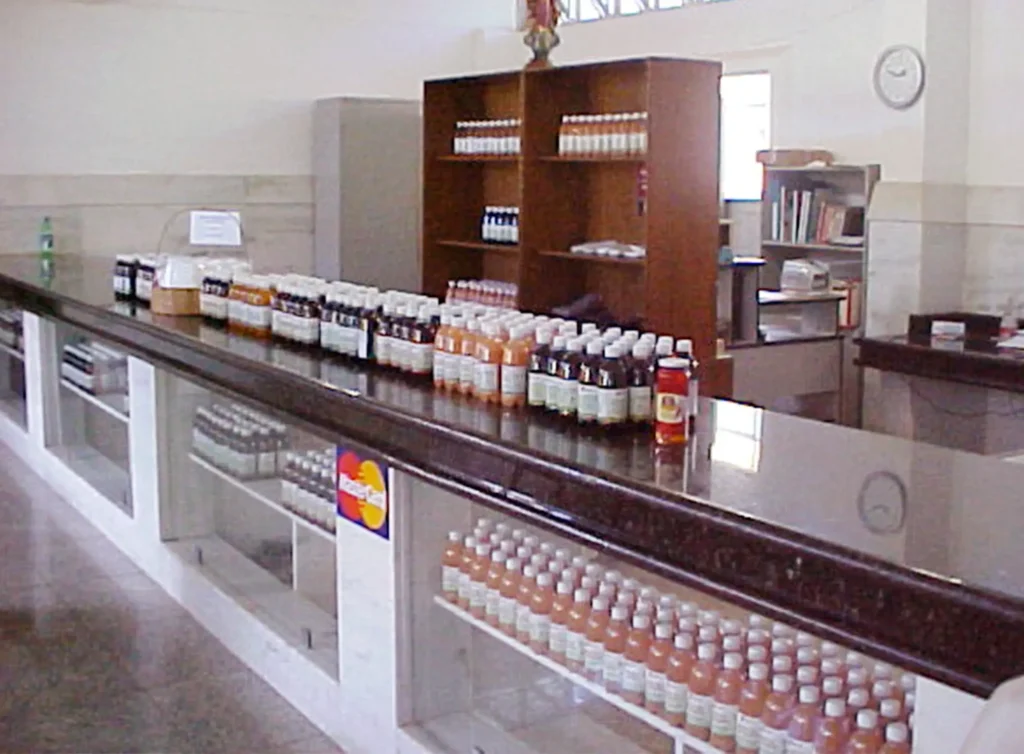 Interior de uma loja ou farmácia com diversos produtos naturais para tratar problemas de saúde, que eram produzidos e comercializados na Fundação Cima’s. A imagem mostra um balcão longo com várias garrafas de produtos naturais, de diferentes tamanhos e cores dispostas em cima. Abaixo do balcão, há prateleiras adicionais com mais garrafas. Uma unidade de prateleiras de madeira fica contra a parede ao fundo, segurando ainda mais garrafas e alguns livros ou caixas à direita.